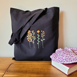 K&ouml;nnte beinhalten: Schwarze Stofftasche mit einem gestickten Blumendesign in Orange und Gelb. Das Design zeigt vier stilisierte Blumen mit Stielen und Bl&auml;ttern.