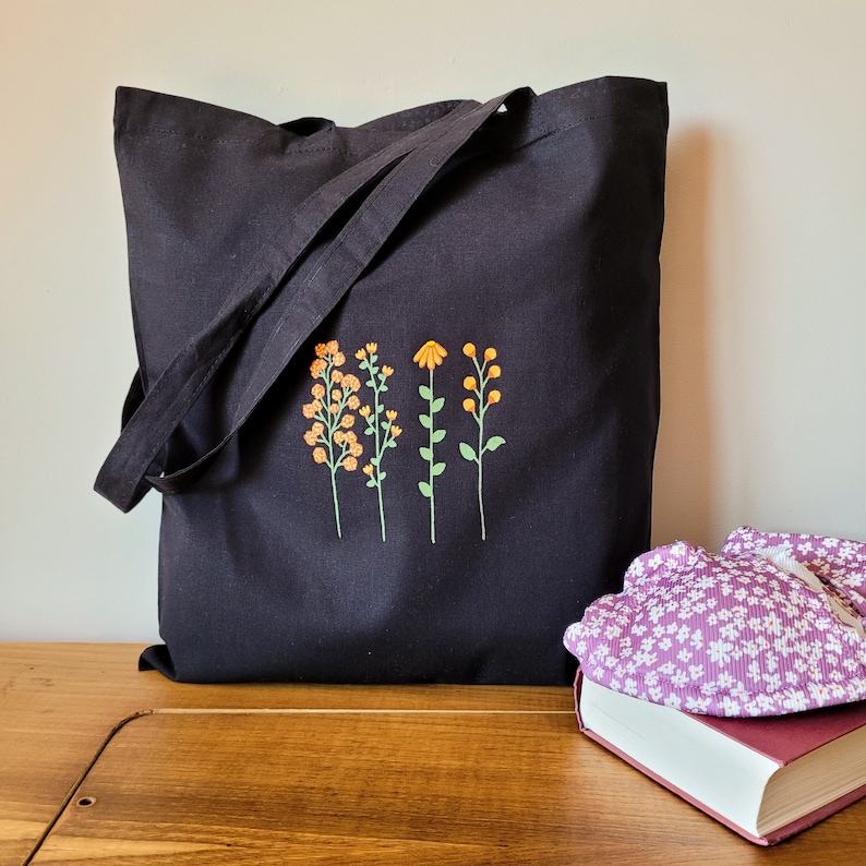 K&ouml;nnte beinhalten: Schwarze Stofftasche mit einem gestickten Blumendesign in Orange und Gelb. Das Design zeigt vier stilisierte Blumen mit Stielen und Bl&auml;ttern.