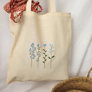 Peut inclure: Un sac fourre-tout beige avec un motif de fleurs bleues et de tiges vertes brod&eacute;es sur le devant.