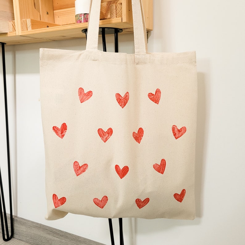 May include: A white canvas tote bag with red heart designs printed on it. The bag has two handles and is made of a natural fabric.