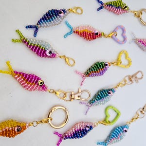 May include: A collection of beaded fish keychains in various colors, including blue, pink, yellow, and green. Each fish has a gold-colored clasp or keyring. The fish are detailed with bead scales and fins, and some have heart-shaped or round attachments.