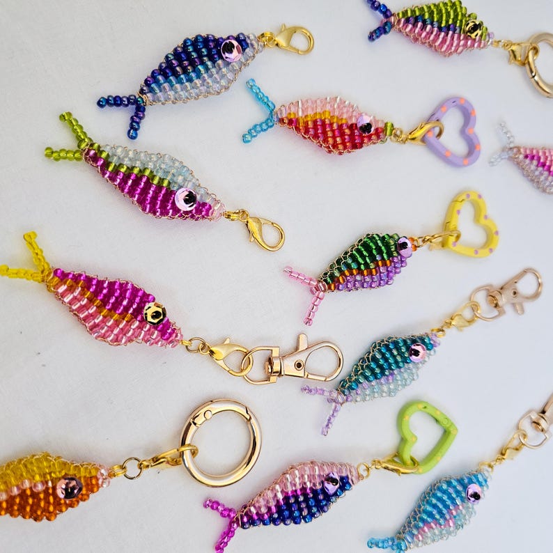 May include: A collection of beaded fish keychains in various colors, including blue, pink, yellow, and green. Each fish has a gold-colored clasp or keyring. The fish are detailed with bead scales and fins, and some have heart-shaped or round attachments.