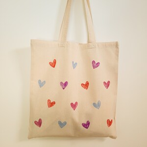 May include: A beige canvas tote bag with a variety of colorful heart designs printed on it. The hearts are in shades of pink, purple, and blue.