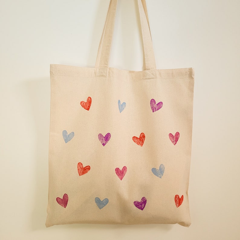 May include: A beige canvas tote bag with a variety of colorful heart designs printed on it. The hearts are in shades of pink, purple, and blue.