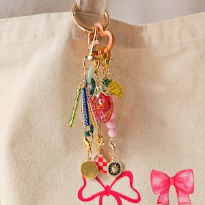 May include: A gold key chain with a variety of colorful charms, including a pink strawberry, green and blue beads, and a gold heart charm. The keychain is hanging from a white canvas tote bag with a pink bow.