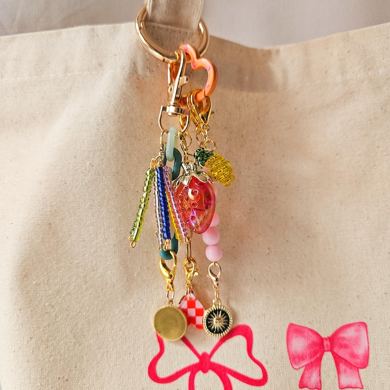 May include: A gold key chain with a variety of colorful charms, including a pink strawberry, green and blue beads, and a gold heart charm. The keychain is hanging from a white canvas tote bag with a pink bow.