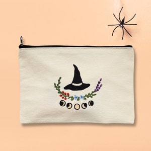 May include: A white canvas zippered pouch with a black embroidered witch hat, flowers, and moons. The pouch is perfect for storing small items like makeup, jewelry, or other accessories.