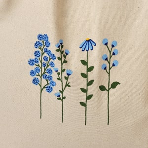 Peut inclure: Un sac fourre-tout avec un motif peint &agrave; la main de quatre fleurs bleues avec des tiges vertes sur un fond de toile naturel.