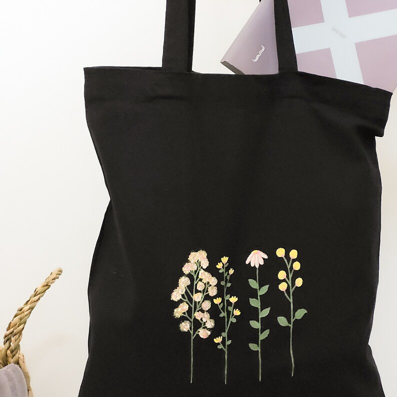 May include: Black canvas tote bag with a floral embroidery design. The embroidery features four different types of flowers in shades of pink, yellow, and green.