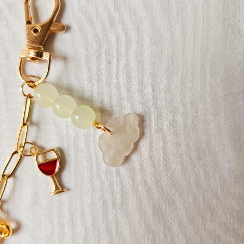 Puede incluir: Llavero dorado con un dije de vino de vidrio, un dije de nube y una cadena de cuentas verde claro.