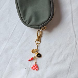May include: A gold-toned charm with various pendants, including a red chili pepper, a black star, a gold coin, and a red and white checkered heart. The charm is attached to a green pouch with a gold zipper.