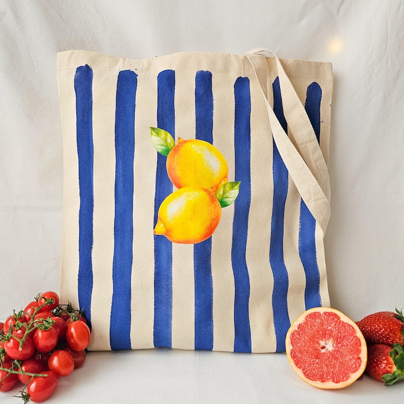 May include: A white canvas tote bag with blue stripes and a painted image of two lemons. The bag has two handles.