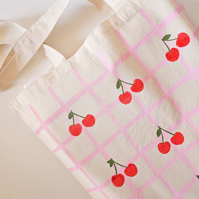 May include: A white canvas tote bag with pink grid pattern and red cherries painted on it.