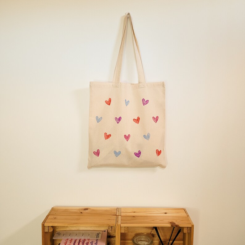 May include: A beige tote bag with a pattern of colorful hearts. The hearts are in shades of pink, red, blue, and purple.