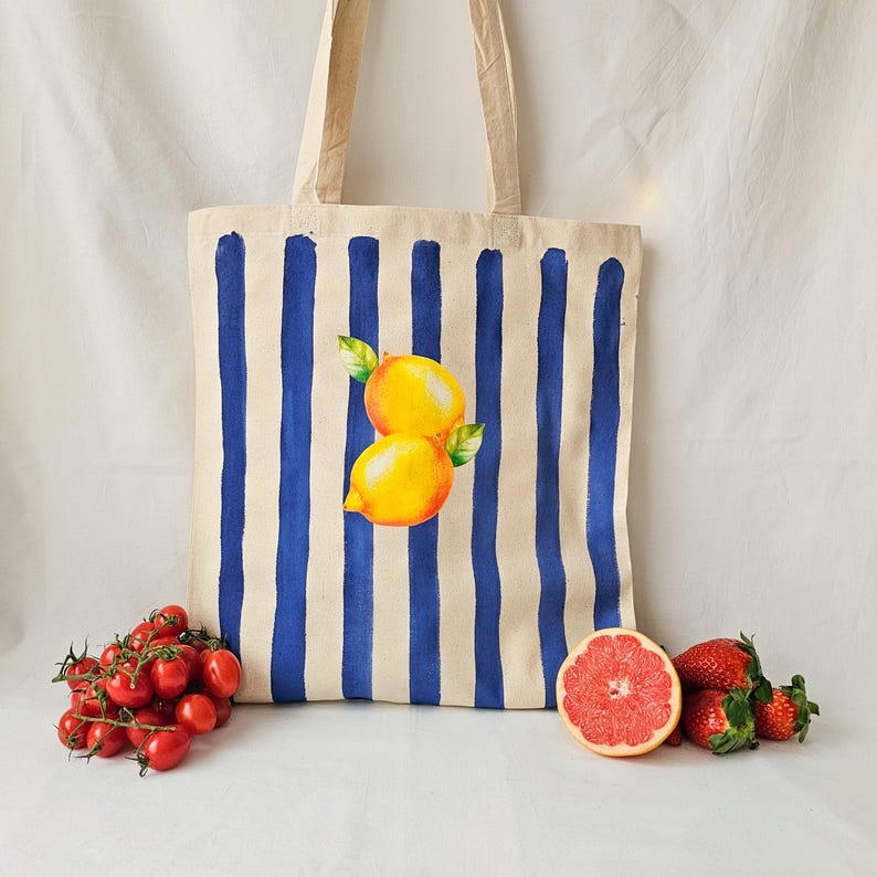 May include: A white canvas tote bag with blue stripes and a painted image of two lemons. The bag has two handles.