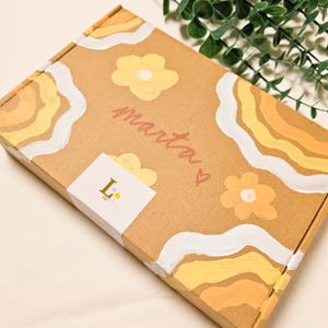 May include: A brown cardboard box decorated with yellow and white floral and wave designs. The name "marta" is written in pink script, with a small heart. A white sticker with the letter "L" and a star is on the box.