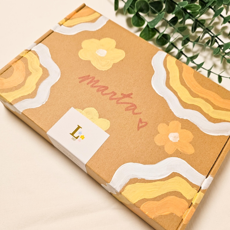 May include: A brown cardboard box decorated with yellow and white floral and wave designs. The name "marta" is written in pink script, with a small heart. A white sticker with the letter "L" and a star is on the box.