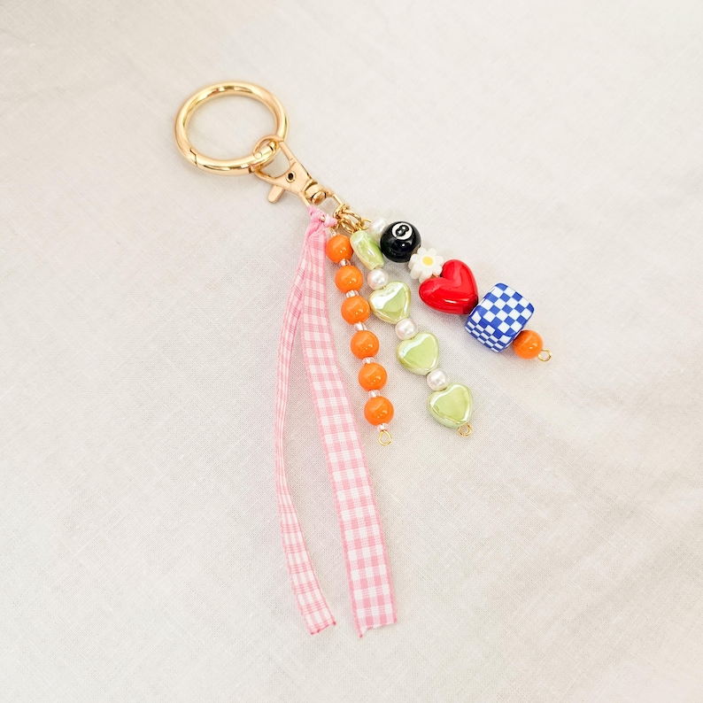 May include: A gold-colored keychain with a lobster clasp, featuring a charm cluster. The cluster includes orange beads, a pink gingham ribbon, green heart beads, a red heart, a blue and white checkered bead, and a black 8-ball bead.