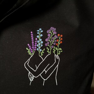 May include: A black tote bag with a white outline of two figures embracing. The figures are holding bouquets of purple, blue, and orange flowers.