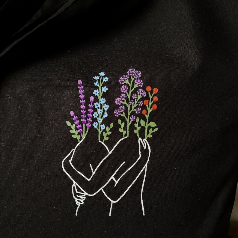 May include: A black tote bag with a white outline of two figures embracing. The figures are holding bouquets of purple, blue, and orange flowers.