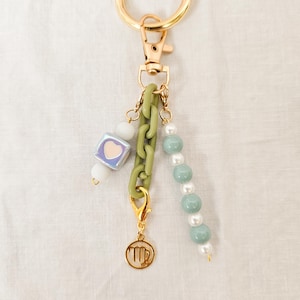 May include: A gold-colored keychain with various charms. The charms include a green chain, a cube with a heart, white beads, and a strand of blue-green beads. A gold-colored charm with a zodiac symbol hangs from the bottom.