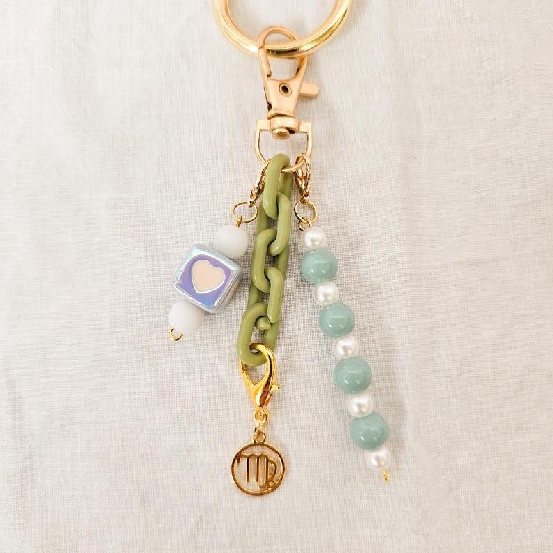 May include: A gold-colored keychain with various charms. The charms include a green chain, a cube with a heart, white beads, and a strand of blue-green beads. A gold-colored charm with a zodiac symbol hangs from the bottom.