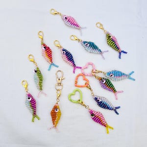 May include: A collection of beaded fish keychains in various colors, including pink, blue, green, and orange. Each fish has a gold-colored clasp and a small tail fin. Some keychains feature heart-shaped or round clasps.