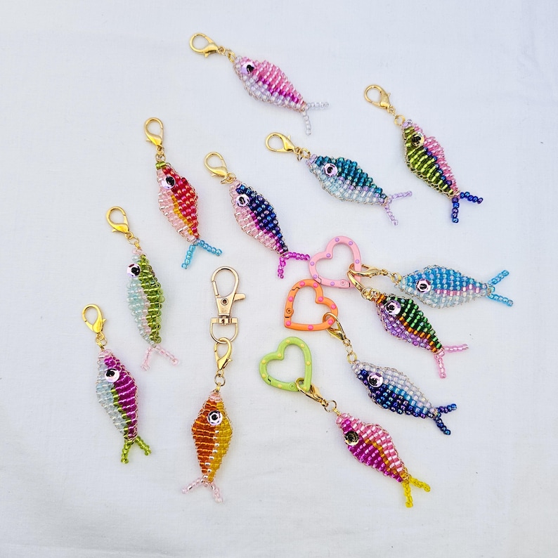 May include: A collection of beaded fish keychains in various colors, including pink, blue, green, and orange. Each fish has a gold-colored clasp and a small tail fin. Some keychains feature heart-shaped or round clasps.