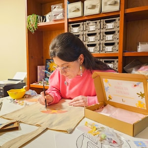 May include: A person painting a design on a beige tote bag with a paintbrush. A cardboard box with the text "KIT PINTA TU TOTE BAG" is open on the table. The person is wearing a pink sweatshirt and glasses. Paint supplies are nearby.