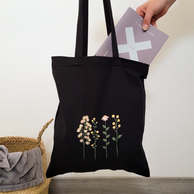 K&ouml;nnte beinhalten: Schwarze Tragetasche mit gestickten Blumen in Rosa-, Gelb- und Gr&uuml;nt&ouml;nen. Die Tasche hat ein Buch mit dem Text "el secreto a vivir" und "know" darin.
