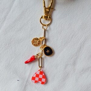 May include: Gold-tone keychain with multiple charms. Charms include a red chili pepper, a black oval with a gold star, a gold compass, and a pink and red checkered heart. The keychain has a lobster clasp.