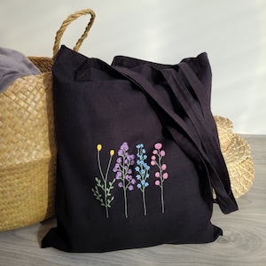 K&ouml;nnte beinhalten: Schwarze Stofftasche mit gesticktem Blumenmuster. Die Blumen sind in den Farben Lila, Blau, Rosa und Gelb.
