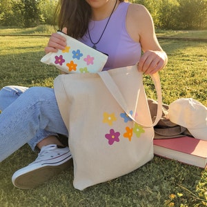 Peut inclure: Un sac fourre-tout en toile blanche avec des motifs de fleurs color&eacute;es. Le sac a une longue sangle et est tenu par une personne portant un jean et un d&eacute;bardeur violet.