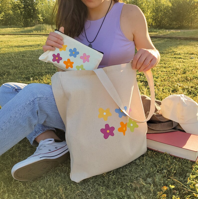 Peut inclure: Un sac fourre-tout en toile blanche avec des motifs de fleurs color&eacute;es. Le sac a une longue sangle et est tenu par une personne portant un jean et un d&eacute;bardeur violet.