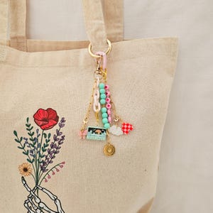 May include: A decorative keychain with multiple charms, including a cassette tape, butterfly, and heart, hangs from a gold-colored clasp. The keychain is attached to a beige tote bag with a floral design.
