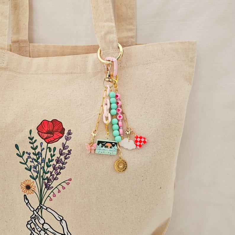 May include: A decorative keychain with multiple charms, including a cassette tape, butterfly, and heart, hangs from a gold-colored clasp. The keychain is attached to a beige tote bag with a floral design.