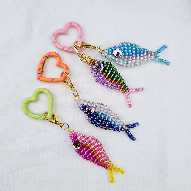 May include: Four beaded fish keychains in various colors: blue, green, purple, and pink. Each fish has a different color scheme and is attached to a heart-shaped clasp. The keychains are on a white background.