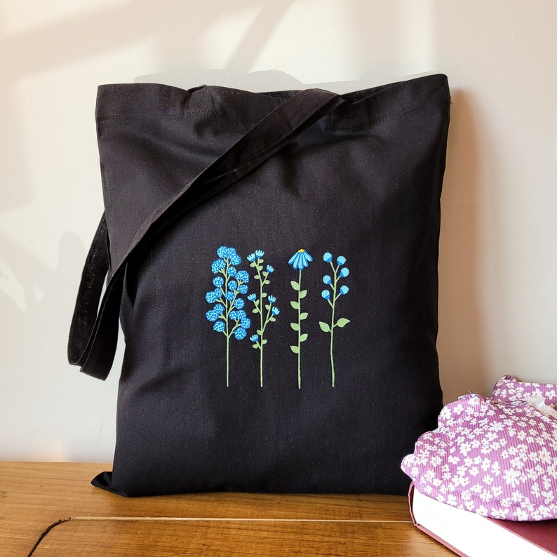 May include: Black tote bag with a design of four blue flowers embroidered on the front. The flowers are stylized and have green stems.
