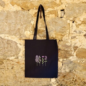 May include: Black tote bag with a floral design embroidered on the front. The flowers are purple, pink, and green.
