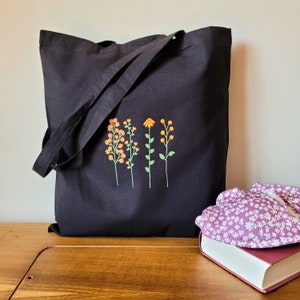 May include: Black canvas tote bag with a floral design embroidered on the front. The design features four stylised flowers in shades of orange and yellow.