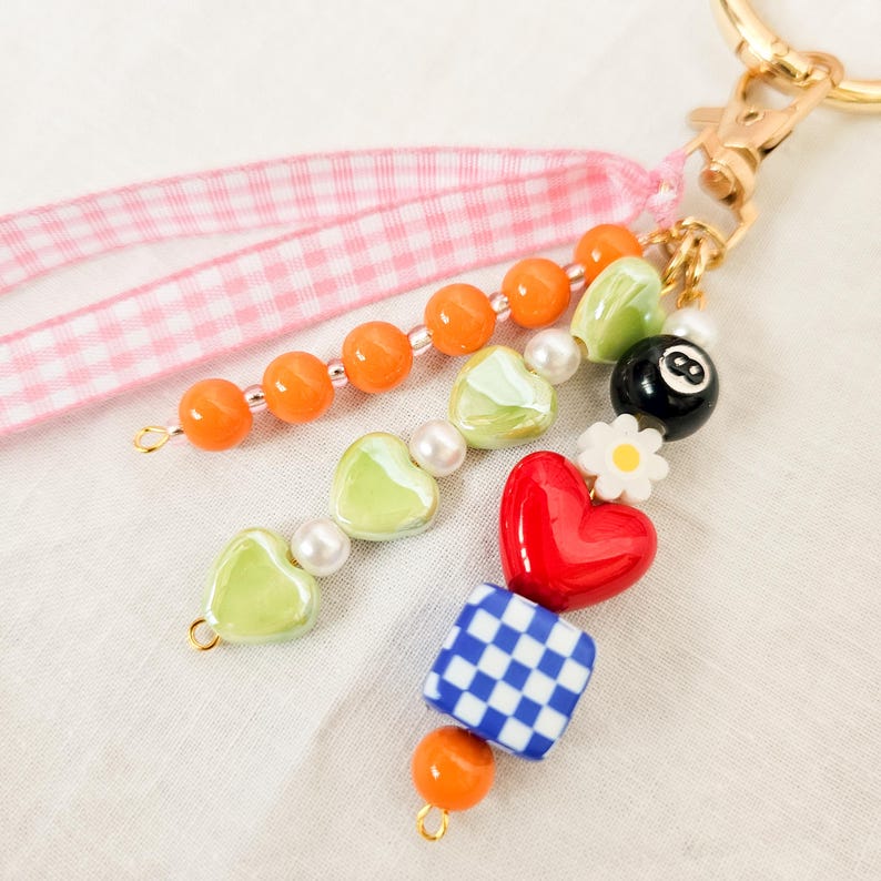 May include: A colorful beaded keychain with a gold clasp. The keychain features orange beads, green heart-shaped beads, a red heart, a blue and white checkered bead, and a black 8-ball bead. A pink gingham ribbon is attached.