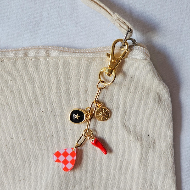 May include: A gold-colored keychain with multiple charms, including a black and gold star, a gold sunburst, a red chili pepper, and a red and white checkered heart. The keychain is attached to a beige canvas bag with a gold clasp.