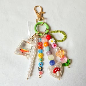 Puede incluir: Un llavero colorido con un cierre dorado y un anillo verde. Cuenta con una pequeña bolsa de plástico con un pez, una cadena de perlas y una cadena de cuentas con varias cuentas de colores, un corazón con un hongo y una flor.