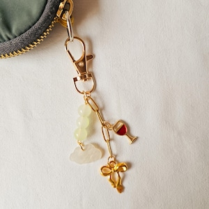 Puede incluir: Un llavero dorado con un dije de copa de vino, una nube, un lazo y tres cuentas verdes.