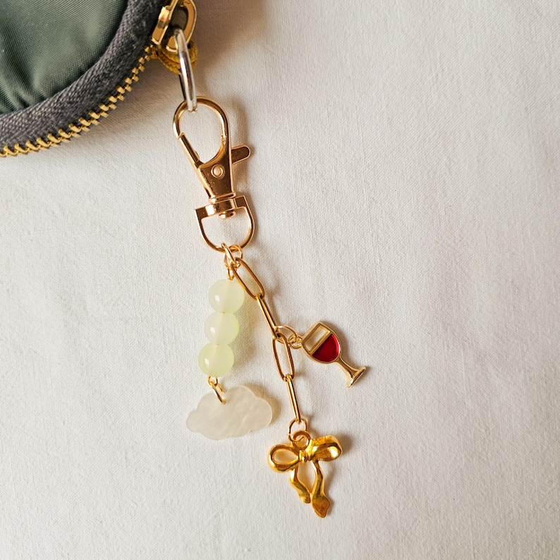 Puede incluir: Un llavero dorado con un dije de copa de vino, una nube, un lazo y tres cuentas verdes.