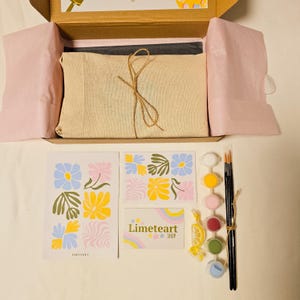 May include: An open cardboard box reveals a paint-by-numbers kit. Inside, a canvas is wrapped in beige fabric and tied with twine. The kit includes paint pots, brushes, and floral-themed art prints. The brand name "Limeteart SHOP" is visible.
