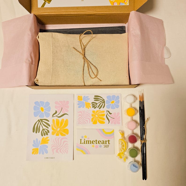 May include: An open cardboard box reveals a paint-by-numbers kit. Inside, a canvas is wrapped in beige fabric and tied with twine. The kit includes paint pots, brushes, and floral-themed art prints. The brand name "Limeteart SHOP" is visible.