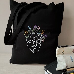 K&ouml;nnte beinhalten: Schwarze Canvas-Tragetasche mit einem wei&szlig;en gestickten Design eines Herzens mit floralen Akzenten. Das Herz ist von bunten Blumen und Bl&auml;ttern umgeben.