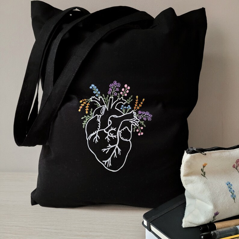 K&ouml;nnte beinhalten: Schwarze Canvas-Tragetasche mit einem wei&szlig;en gestickten Design eines Herzens mit floralen Akzenten. Das Herz ist von bunten Blumen und Bl&auml;ttern umgeben.