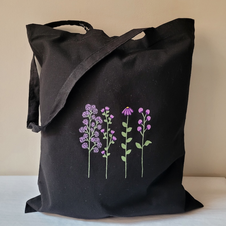 May include: Black canvas tote bag featuring an embroidered design of purple flowers. The flowers are arranged in a row and have green stems.